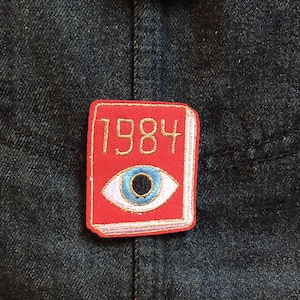 May include: A red patch featuring the text "1984" and an eye design. The patch has gold stitching and a white border. The eye is blue with a black pupil. The patch is on a dark blue denim jacket.