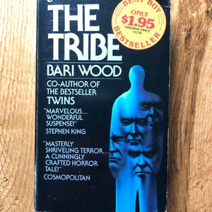 May include: A paperback book cover with a blue background and a white title that reads "The Tribe" by Bari Wood. The book is a bestseller and is co-authored by Bari Wood. The cover features a blue silhouette of a person with multiple faces. The book is a horror novel and has positive reviews from Stephen King and Cosmopolitan.