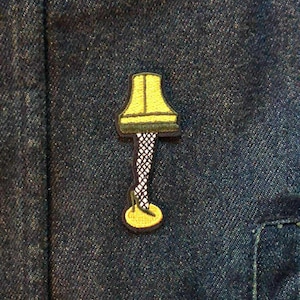 Peut inclure: Écusson brodé représentant une lampe jaune avec un contour noir et une base en forme de jambe. L'écusson est posé sur un fond en denim bleu foncé, avec des coutures visibles et un détail de poche. Un accessoire amusant.