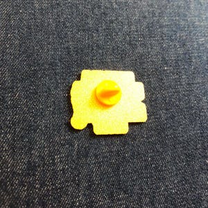 May include: A yellow metal pin back with a textured surface and a central yellow rubber clutch fastener. The pin back is irregular in shape and features a flat, granular finish on the reverse side, showing the mechanism used to attach the item.