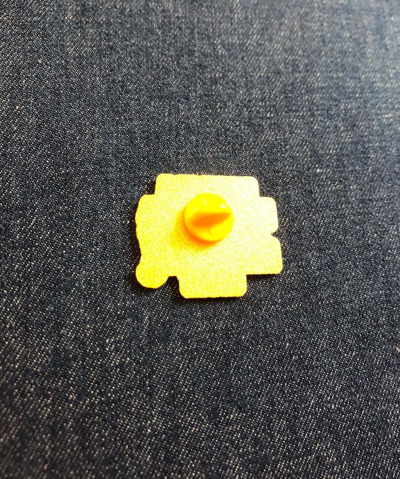 May include: A yellow metal pin back with a textured surface and a central yellow rubber clutch fastener. The pin back is irregular in shape and features a flat, granular finish on the reverse side, showing the mechanism used to attach the item.