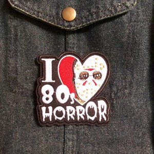 May include: Embroidered patch with the text "I 80s HORROR" in white lettering on a black border. The patch features a heart-shaped design with a red and white mask. The patch is attached to a dark denim jacket.