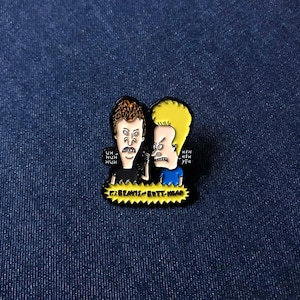 May include: Enamel pin depicting Beavis and Butt-Head. Beavis is in a black shirt, Butt-Head in blue. The pin includes the text "BEAVIS BUTT-HEAD" on a yellow banner, along with their signature phrases.