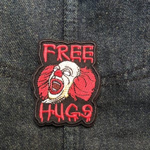 May include: Embroidered patch showing a scary clown with red hair and a white face, with the words "FREE HUGS" in red lettering. The patch is sewn onto a dark blue denim fabric.