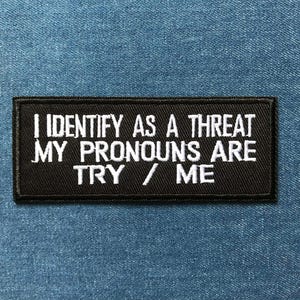 Peut inclure: Un écusson rectangulaire noir avec le texte brodé en blanc : "I IDENTIFY AS A THREAT MY PRONOUNS ARE TRY / ME." L'écusson est posé sur un fond en denim bleu.