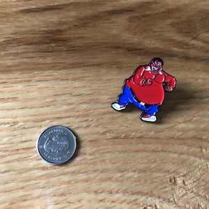 May include: A vibrant enamel pin depicting a cartoon figure in a red top, blue trousers, and white trainers. The pin is on a wooden surface next to a coin. The character is in a running pose.