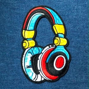 May include: An embroidered patch shaped like a pair of over-ear headphones. The design features a bright blue headband with yellow accents, and ear cups colored in white, black, and red, all outlined with a thick black border for definition.