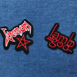 May include: Two embroidered patches with black borders and red lettering. One patch reads "Venom" with a red star. The other patch reads "Lamb of God". Both patches are on a blue denim background.