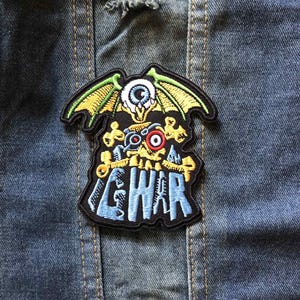 May include: Embroidered patch with a black background featuring a winged eye design, bones, and the word "GWAR" in blue. The patch has green, yellow, red, and white accents, and is set against a denim jacket.