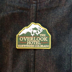 May include: A rectangular patch with a green mountain and white snow design, and the words "THE OVERLOOK HOTEL SIDEWINDER COLORADO" in white and gold lettering. The patch is on a dark blue denim background.