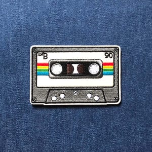 May include: An embroidered patch shaped like a cassette tape with a grey frame, white centre, and a horizontal rainbow stripe. The text "SIDE B" is printed on the top left and "90" on the top right. The design includes two white circular spools.