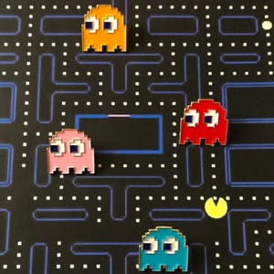 May include: Enamel pins of Pac-Man ghosts in orange, red, pink, and teal. The pins are set against a black background with a Pac-Man game board design. Each ghost has white eyes and a square shape.