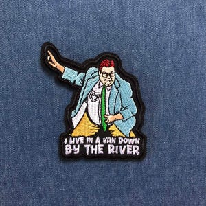May include: Embroidered patch featuring a cartoon man in a light blue blazer, white shirt, and green tie. The patch includes the text "I LIVE IN A VAN DOWN BY THE RIVER" in white letters on a black background.