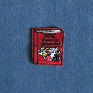 May include: A red embroidered patch in the shape of a book. The book has the words "Handbook for the Recently Deceased" on the cover. The image on the book shows two figures in a scene with a yellow and blue background.