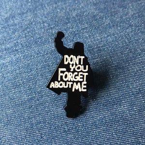 May include: Black enamel pin in the shape of a silhouette with the text "DON'T YOU FORGET ABOUT ME" in white. The pin is on a blue denim background.