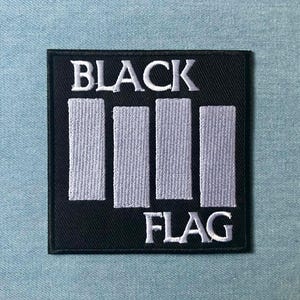 Peut inclure: Un écusson carré noir avec les mots "BLACK" et "FLAG" brodés en blanc. Le patch présente quatre barres verticales blanches. Il s'agit probablement d'un emblème ou d'un logo.