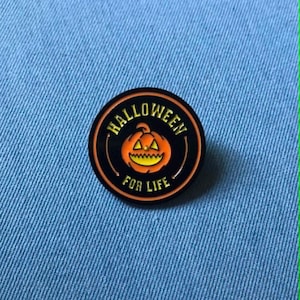 May include: A circular enamel pin featuring a black background with an orange jack-o'-lantern in the centre. The text HALLOWEEN is arched above the pumpkin and FOR LIFE is arched below, both in yellow lettering with a thin orange border.