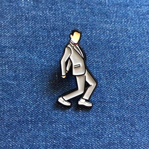Peut inclure: Une épingle en émail grise représentant un homme en costume et cravate, dans une pose de marche stylisée. L'épingle a un contour noir et est posée sur un fond en denim bleu.