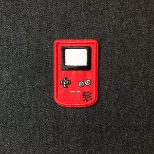 May include: A red embroidered patch depicting a retro handheld games console. The patch features a black screen, directional pad, buttons, and speaker details. The patch is rectangular with rounded corners.