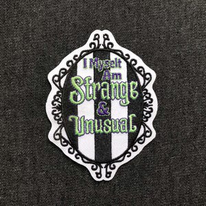 Peut inclure: Écusson brodé avec le texte "I Myself Am Strange & Unusual" en vert et violet, sur un fond à rayures noires et blanches. L'écusson a une bordure noire décorative et un contour blanc.