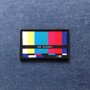 Puede incluir: Un parche rectangular con borde negro que presenta un patrón de prueba colorido de barras verticales en amarillo, azul, cian y rojo. El texto "NO SIGNAL" se muestra en blanco en el centro del parche.