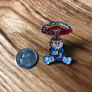 May include: A blue and white enamel pin of a cartoon character with a red and yellow mushroom cloud over their head. The character is wearing a blue suit and has a white collar.
