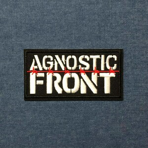 May include: A black rectangular patch with the words "AGNOSTIC FRONT" in white block letters. The text is bisected by a red line with small red accents. The patch is on a blue denim background.