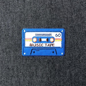 May include: A blue embroidered patch in the shape of a cassette tape. The patch features the words "MUSIC TAPE" and the number "60" in white. The tape has orange, yellow, and red stripes.