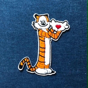May include: An embroidered patch featuring a cartoon tiger with orange fur and black stripes, standing upright with a white belly and paws. The tiger holds a pair of white gloves with a small, bright red heart centred between the hands.