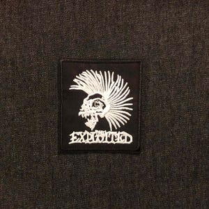 May include: Black square patch with a white embroidered skull wearing a mohawk. The word "EXHIBITED" is embroidered in white below the skull. The patch is set against a dark gray fabric background.