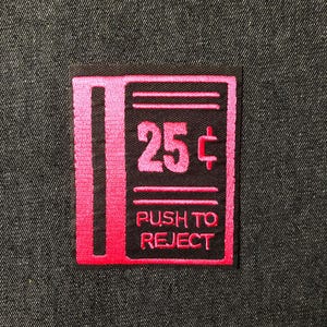 May include: A square, embroidered patch with a black background and pink accents. The patch features the number "25p" and the words "PUSH TO REJECT" in pink. The patch is likely designed to be sewn onto clothing or accessories.