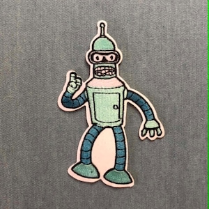 May include: An embroidered patch featuring a cartoon robot with a light green head and torso, dark blue segmented arms and legs, and light green feet. The robot has a rectangular chest door, a wide grin with visible teeth, and one raised finger.