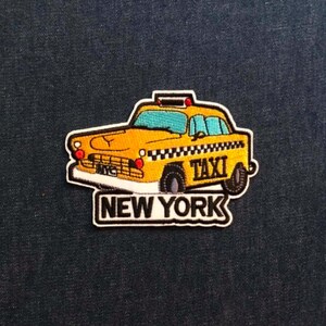 May include: An embroidered patch featuring a yellow New York taxi cab with a black and white checkered stripe along the side. The word TAXI is stitched on the door, and the text NEW YORK is displayed in bold black letters on a white background.