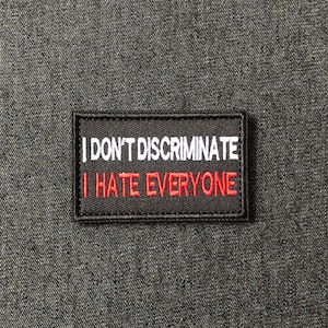 Peut inclure: Un écusson rectangulaire noir avec le texte brodé en blanc « I DON'T DISCRIMINATE » au-dessus du texte brodé en rouge « I HATE EVERYONE ». L'écusson est sur un tissu texturé gris foncé.