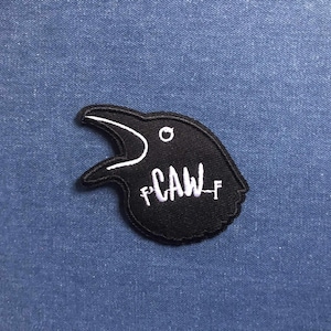 May include: Black crow patch with white embroidered details. The patch features the word "CAW" in white script. The crow's beak is open, and there is a small white circle for the eye. The patch is on a blue background.