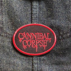 May include: Oval patch with the text "CANNIBAL CORPSE" in red, stitched onto a black background with a red border. The patch is affixed to a dark blue denim jacket.