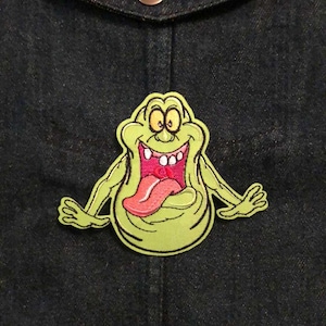 May include: A green cartoon character patch with a pink tongue and white teeth is sewn onto a dark blue denim jacket. The patch has a yellow outline and is centered on the jacket.