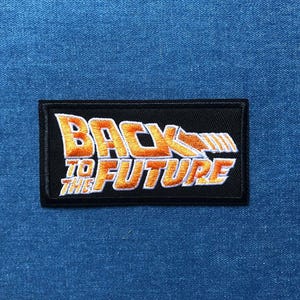 May include: Embroidered patch featuring the text "BACK TO THE FUTURE" in orange and white, with a stylized arrow and lines design. The patch is rectangular with a black border, set against a blue fabric background.