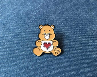 Può includere: Una spilla di Care Bear color tan con un cuore rosso sulla pancia. La spilla ha un contorno nero ed è posta su uno sfondo di denim blu. L'orso è in posizione seduta con le braccia e le gambe distese.