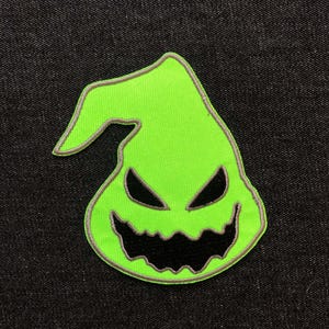 May include: A neon green patch featuring a stylized, grinning face with black eyes and mouth. The patch has a gray border and is set against a dark denim background. The design is reminiscent of a cartoon character.
