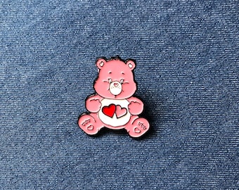 Puede incluir: Pin de esmalte rosa con un oso de dibujos animados con dos corazones rojos en la barriga. El oso tiene un hocico blanco y un contorno negro. El pin está sobre un fondo de mezclilla azul.