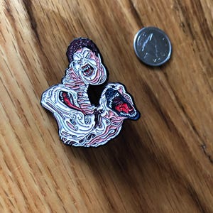 May include: A horror-themed enamel pin featuring a grotesque, screaming figure with exposed flesh and a gaping mouth. The pin has a white, pink, and red color scheme with black outlines. A silver coin is next to the pin.