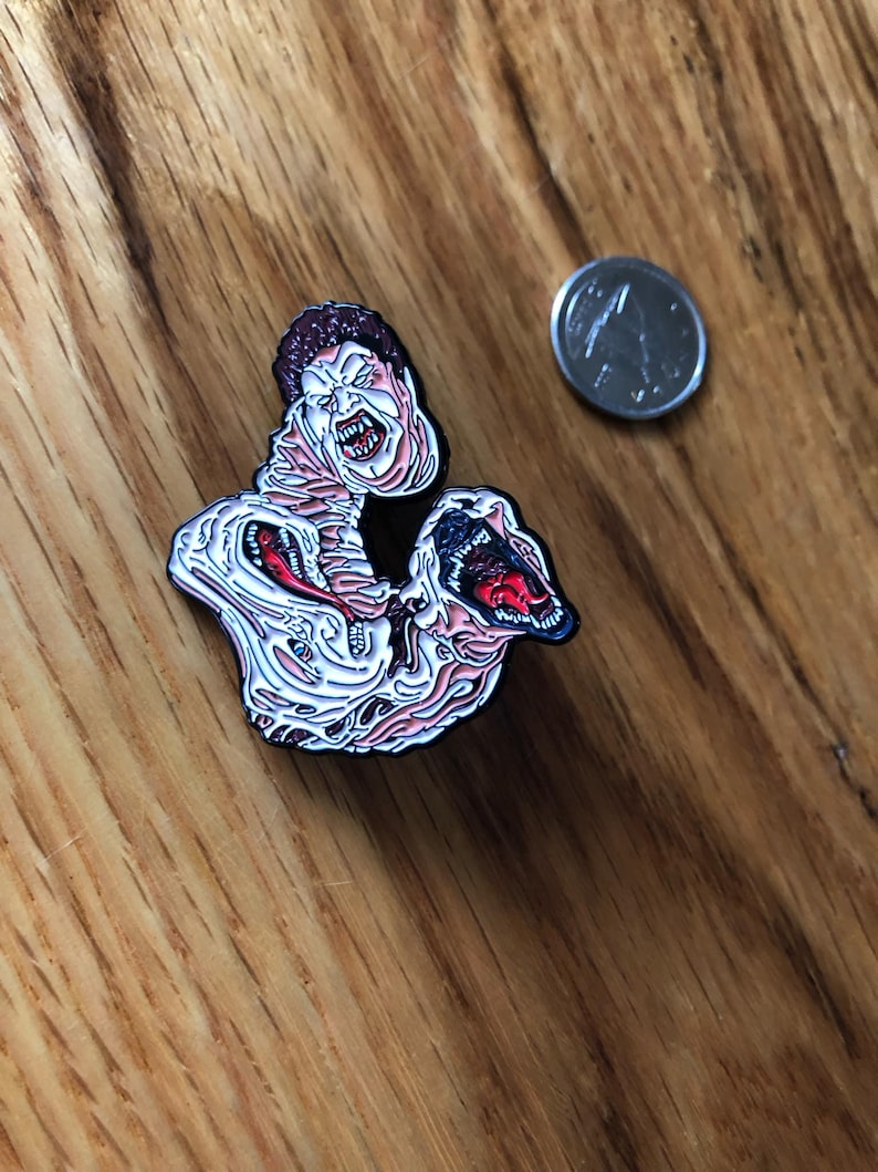 May include: A horror-themed enamel pin featuring a grotesque, screaming figure with exposed flesh and a gaping mouth. The pin has a white, pink, and red color scheme with black outlines. A silver coin is next to the pin.