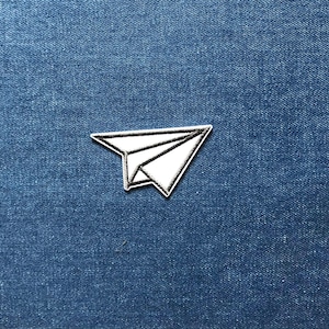May include: A white paper aeroplane patch with a black outline and details, set against a blue denim background. The patch is a simple, geometric design, ideal for adding a touch of whimsy to clothing or accessories.