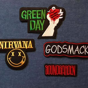 May include: A selection of embroidered patches displaying band logos and graphics. Patches include Green Day, Nirvana, Godsmack, and Soundgarden. The Nirvana patch features a smiley face design. The patches are black with various colours.