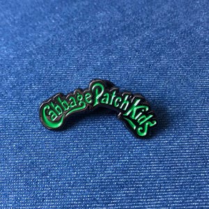 May include: A Cabbage Patch Kids enamel pin with a black outline and green lettering. The pin is curved and set against a blue denim background. The pin is a collectible item.