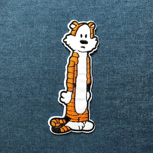 May include: Embroidered patch of a cartoon tiger character in orange, black, and white. The patch has a white border and is set against a blue denim background. The tiger is standing with a thoughtful expression.