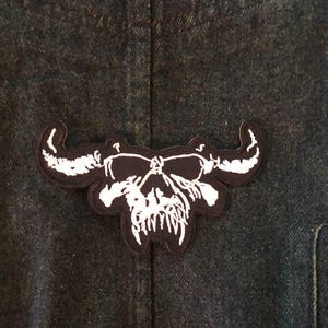 May include: A black and white skull patch with horns, attached to a dark denim jacket. The patch features a detailed skull design with prominent teeth and curved horns, creating a bold, edgy aesthetic.