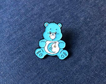 Può includere: Spilla in smalto azzurro a forma di orso, con pancia bianca con un design a mezzaluna e stella. L'orso ha un contorno nero ed è posto su uno sfondo di denim blu. La spilla è un oggetto da collezione.