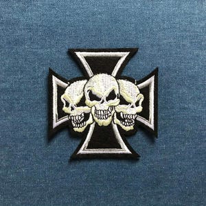 May include: Embroidered patch featuring a black and white design of a Maltese cross with three skulls. The skulls have detailed bone structures and are embroidered with white thread. The patch is on a blue denim background.